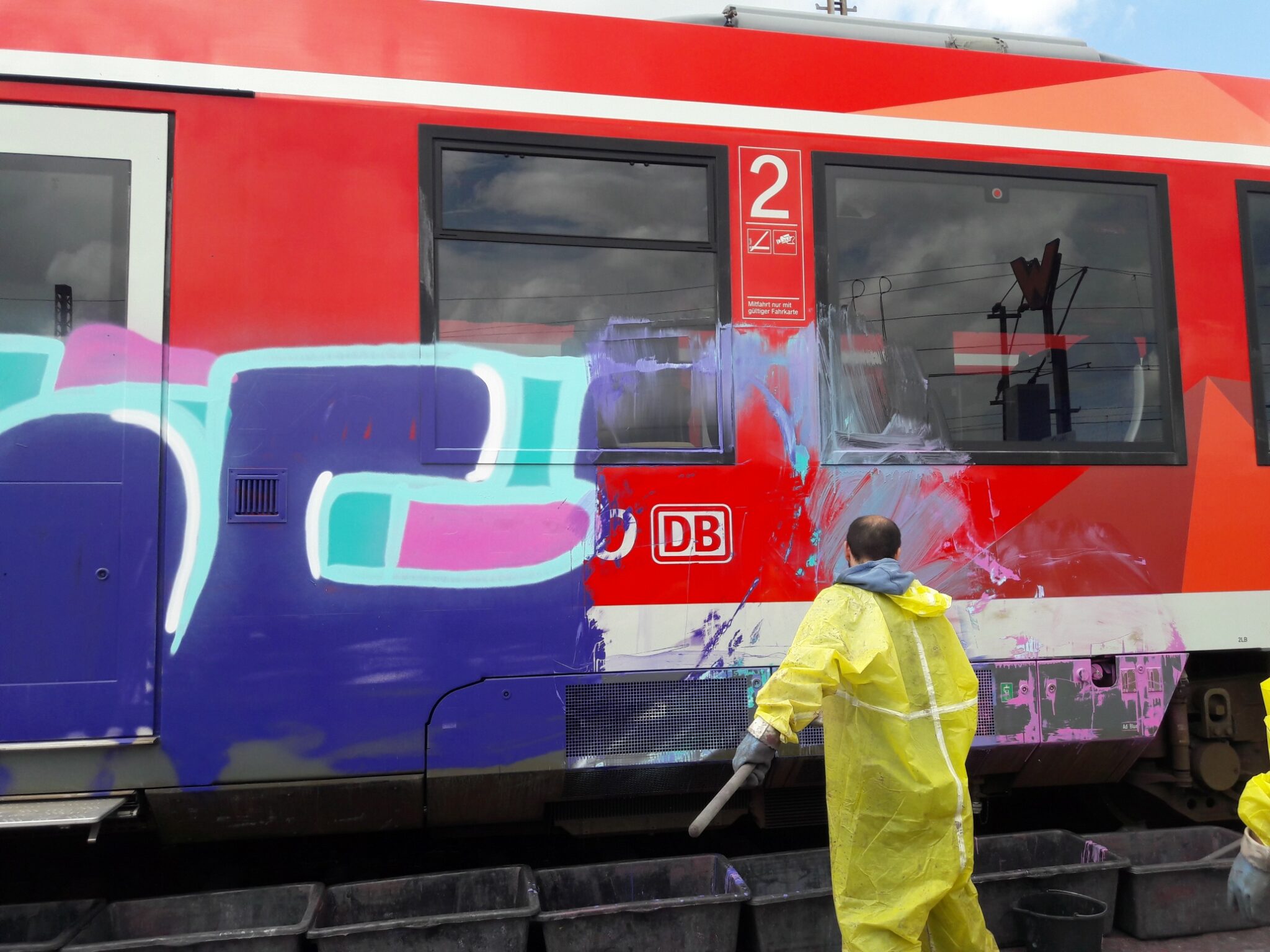 Ein Mann in einem gelben Schutzanzug entfernt Graffiti mit Graffiti-Ex von einem roten Zugwagen.